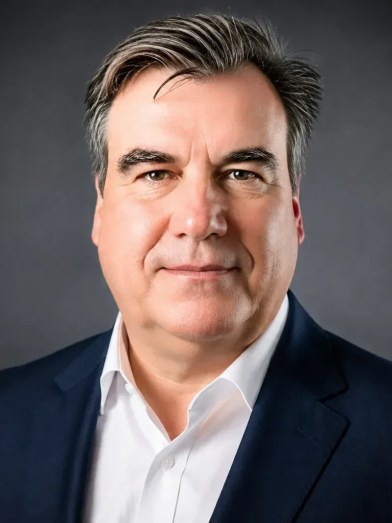 A man wearing a dark blue suit and white shirt against a dark background.